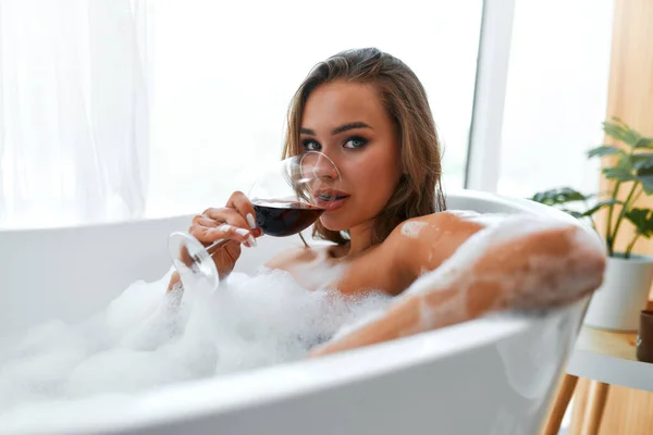 美しいです情熱的なセクシー女性取りますリラックスバスとともに泡と飲みます赤ワインで白いバスルームに窓辺 — ストック写真