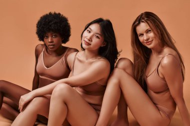 Bej iç çamaşırlı çok ırklı genç kadınlar bej arka planda izole edilmiş. Stüdyoda poz veren Afro-Amerikalı, Kafkas ve Asyalı kadınlar. Yüz bakımı, kozmetoloji kavramı.