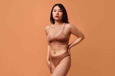 Bej rengi iç çamaşırlı, bej arkaplanda poz veren Asyalı genç bir kadının portresi. Cilt bakımı, kozmetoloji ve kaplıca tedavisi kavramı.