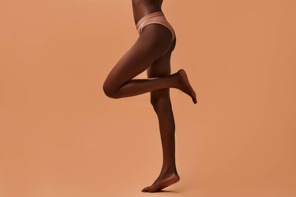Cropped portrait of the legs of an African-American slim woman in beige lingerie isolated on a beige background. Concept of skin care, cosmetology and spa treatments.