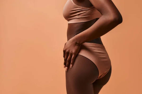 Cropped image of African-American skinny woman in beige underwear isolated on beige background. Cosmetology, spa treatments and body care.