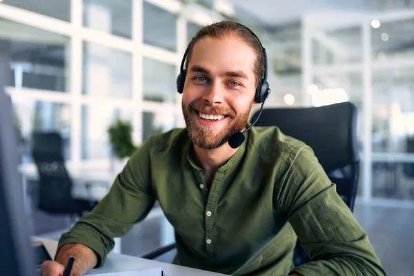 Technical support operator working with headset in office. Smiling handsome man working as call centre operator, speaking to customer. Happy businessman working remotely while doing video conference.