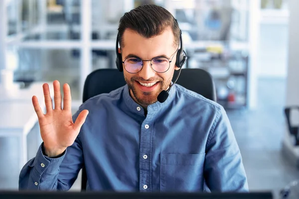 Technical support operator working with headset in office. Smiling handsome man working as call centre operator, speaking to customer. Happy businessman working remotely while doing video conference.