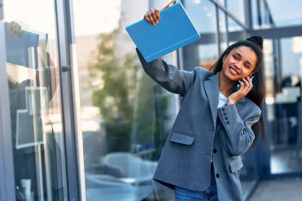 Young stylish woman in a suit talking on the phone and holding a notebook while standing at the entrance to a business center or office building.
