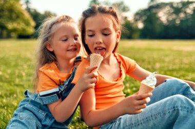 Güneşli bir günde iki kız kardeş parkta çimlerde otururken dondurma yer. İki çocuğun kızı, çayırdaki çimlerin üzerinde oturmuş, tatlı yiyor ve dinleniyorlar..