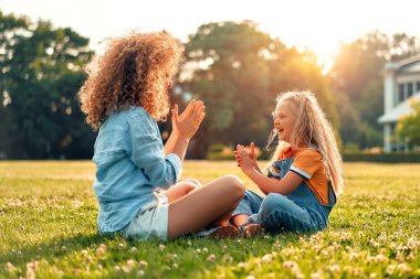 Anne, küçük bir çocuk, kız, oyun oynuyor ve ellerini çırpıyor, güneşli bir günde parkta çimlerin üzerinde çimlerde oturuyor, eğleniyor ve doğanın tadını çıkarıyor..