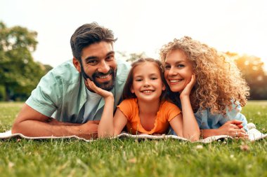 Mutlu, genç bir aile babası, anne ve kızı güneşli bir günde parkta çimlerin üzerinde uzanıyor, birlikte eğleniyor ve tatil günlerinde dinleniyorlar..