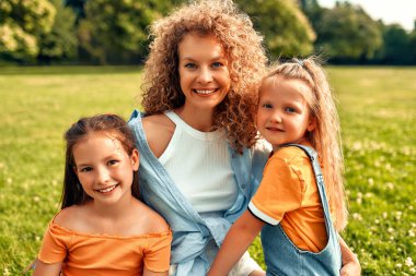 Mutlu genç anne parkta çayırda iki sevgili kızıyla oturuyor, sarılıyor ve onları öpüyor, eğleniyor ve güneşli bir haftasonunda oynuyor..