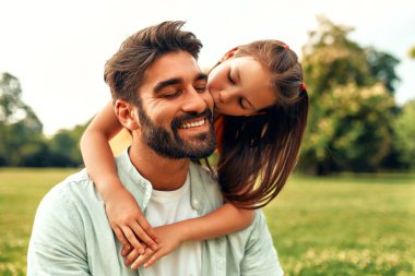 Şirin küçük kız çocuğu, sıcak ve güneşli bir haftasonunda parkta çayırda oturmuş, babasını nazikçe kucaklıyor ve öpüyor..