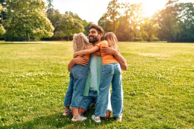 Mutlu genç baba, sıcak güneşli bir günde parkta çayırda oturup eğlenirken iki sevimli kızına sarılıyor..