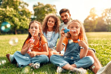 Mutlu bir aile babası, anne ve iki kızı parktaki çimlerin üzerinde sabun köpüğü üflüyor, güneşli bir günde birlikte oynuyor ve eğleniyorlar..
