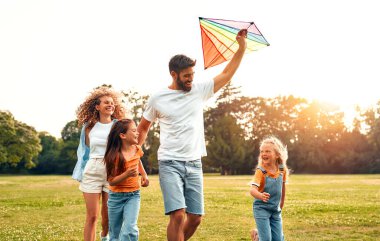 Mutlu bir aile babası, anne ve iki kızı parktaki çayırda renkli bir uçurtmayla koşup oynuyorlar, güneşli bir hafta sonunda eğleniyorlar..