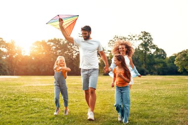 Mutlu bir aile babası, anne ve iki kızı parktaki çayırda renkli bir uçurtmayla koşup oynuyorlar, güneşli bir hafta sonunda eğleniyorlar..