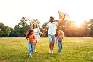 Mutlu bir aile babası, anne ve iki kızı parktaki çayırda renkli bir uçurtmayla koşup oynuyorlar, güneşli bir hafta sonunda eğleniyorlar..