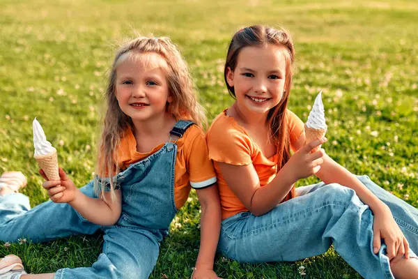 Two sisters eat ice cream while sitting on the lawn in the park on a sunny warm day. Two children\'s girls playing and eating sweets, sitting on the grass in the meadow and resting.