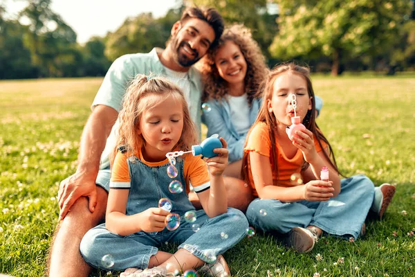 Mutlu bir aile babası, anne ve iki kızı parktaki çimlerin üzerinde sabun köpüğü üflüyor, güneşli bir günde birlikte oynuyor ve eğleniyorlar..