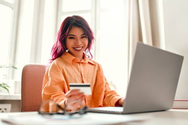 Parlak pembe saç telleri olan Asyalı genç bir kız elinde kredi kartıyla evde dizüstü bilgisayarla bir masada oturuyor. Online alışveriş ve bankacılık.