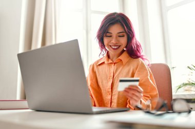 Parlak pembe saç telleri olan Asyalı genç bir kız elinde kredi kartıyla evde dizüstü bilgisayarla bir masada oturuyor. Online alışveriş ve bankacılık.
