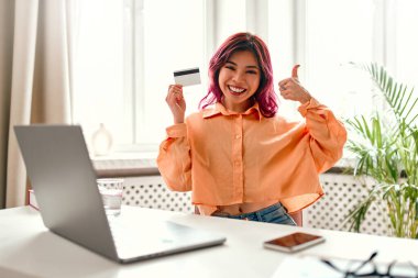 Parlak pembe saç telleri olan Asyalı genç bir kız evinde dizüstü bilgisayarla bir masada oturuyor, elinde bir kredi kartı tutuyor ve onayını veriyor. Online alışveriş ve bankacılık.