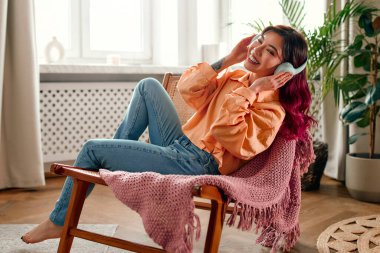 Genç, parlak Asyalı bir kadının portresi. Pembe saç telleriyle müzik dinliyor ya da kulaklıkla kulaklık takıyor. Oturma odasında dinleniyor..