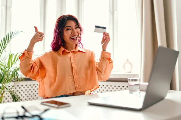 Parlak pembe saç telleri olan Asyalı genç bir kız evinde dizüstü bilgisayarla bir masada oturuyor, elinde bir kredi kartı tutuyor ve onayını veriyor. Online alışveriş ve bankacılık.
