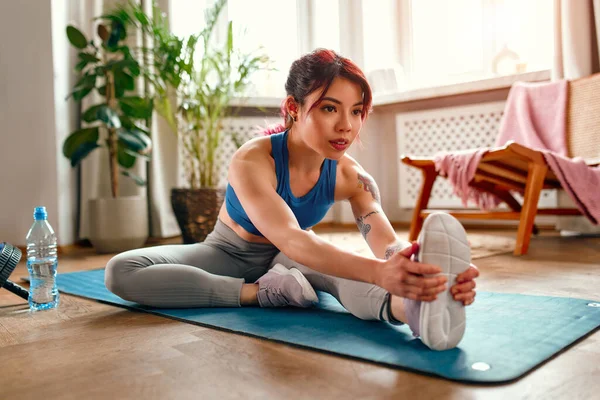 年轻的亚洲女人 身上有纹身 穿着运动服 在客厅的橡皮垫上锻炼 做伸展运动 瑜伽和积极的生活方式 — 图库照片