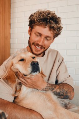 Oturma odasında sevgili köpeğiyle kanepede oturan genç ve yakışıklı bir kıvırcık adam..