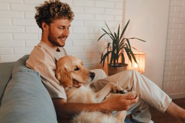 Oturma odasında sevgili köpeğiyle kanepede oturan genç ve yakışıklı bir kıvırcık adam..