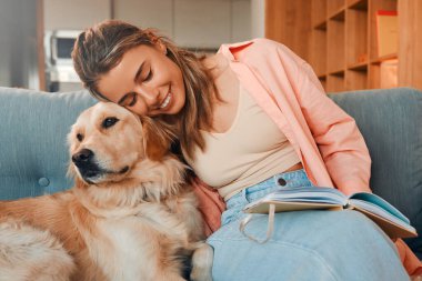Güzel genç bir kadın, kanepede sevgili köpeğiyle birlikte rahat bir evde oturuyor ve kitap okuyor..