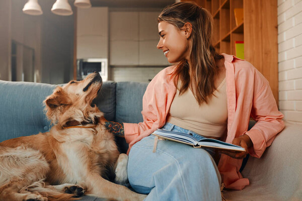 Beautiful young woman sitting on the sofa with her beloved dog in the living room in a cozy home and reading a book.