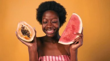 Afro saçlı, elinde papaya ve ellerinde karpuz olan turuncu arka planda yaşayan Afro-Amerikalı genç bir kadın. Cilt bakımı ve kaplıca tedavisi.
