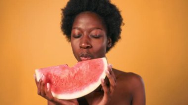 Afro saçlı Afrikalı genç bir kadın turuncu arka planda izole edilmiş lezzetli bir karpuz ısırığı alıyor. Cilt bakımı. Sağlıklı yaşam tarzı.