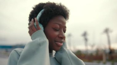 Güzel bir Afro-Amerikalı kadın dışarıda bir kazakla duruyor ve kulaklıkla müzik dinliyor, gözlerini kapıyor ve eğleniyor..