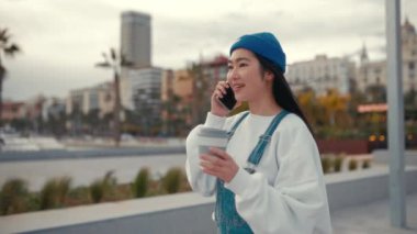 Süveter ve şapka giyen, kahve içen ve caddede yürürken telefonla konuşan şık Asyalı kadın..