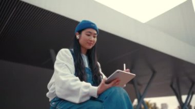 Sokakta tabletli, şapkalı tatlı Asyalı genç bir kadın. Grafik tableti çiziyorum. Serbest ve çevrimiçi kazanç.