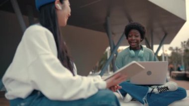 Genç Afro-Amerikalı ve Asyalı kadınlar dışarıda dizüstü bilgisayar ve tabletle yan yana oturuyorlar. Serbest ve çevrimiçi kazanç.