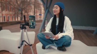 Genç, sevimli Asyalı kadın blogcu, tabletli, akıllı telefondan kanalı için video kaydediyor. Serbest ve çevrimiçi kazanç.