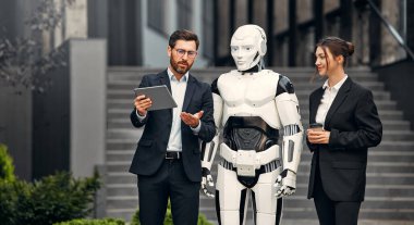 Takım elbiseli genç adam ve kadın iş ortakları ve tabletleri bir robotla yan yana duruyor ve bir binanın arka planında yapılan çalışmaları tartışıyorlar. Yapay zekayla gelecek.