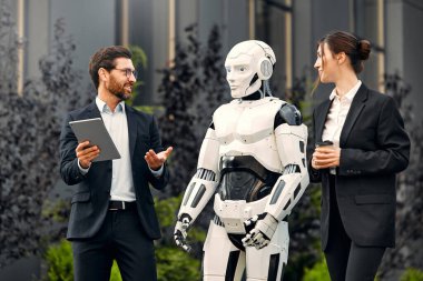 Takım elbiseli genç adam ve kadın iş ortakları ve tabletleri bir robotla yan yana duruyor ve bir binanın arka planında yapılan çalışmaları tartışıyorlar. Yapay zekayla gelecek.