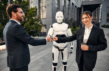 Takım elbiseli genç adam ve kadın iş ortakları bir robotla yan yana duruyor ve yumruklarını birbirine bağlıyorlar. Yapay zekayla gelecek.