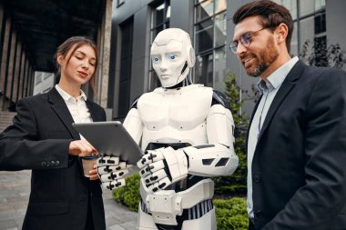 Takım elbiseli genç adam ve kadın iş ortakları ve tabletleri bir robotla yan yana duruyor ve bir binanın arka planında yapılan çalışmaları tartışıyorlar. Yapay zekayla gelecek.