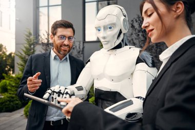 Takım elbiseli genç adam ve kadın iş ortakları ve tabletleri bir robotla yan yana duruyor ve bir binanın arka planında yapılan çalışmaları tartışıyorlar. Yapay zekayla gelecek.