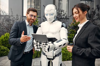 Takım elbiseli genç adam ve kadın iş ortakları ve tabletleri bir robotla yan yana duruyor ve bir binanın arka planında yapılan çalışmaları tartışıyorlar. Yapay zekayla gelecek.
