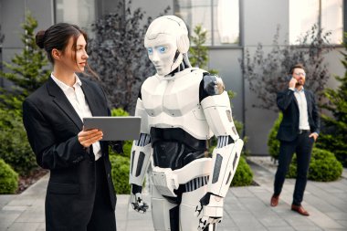 Takım elbiseli genç adam ve kadın iş ortakları ve tabletleri bir robotla yan yana duruyor ve bir binanın arka planında yapılan çalışmaları tartışıyorlar. Yapay zekayla gelecek.