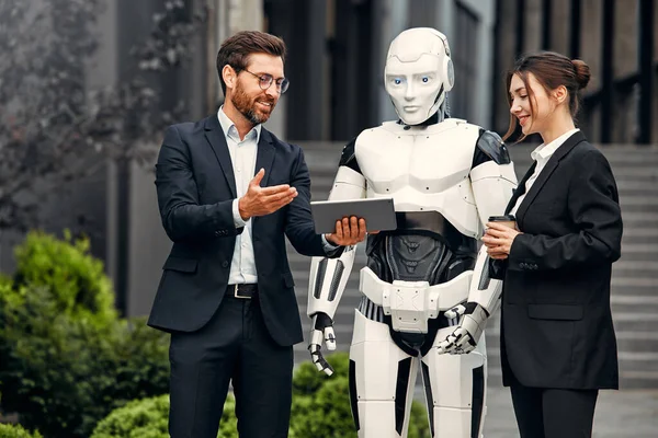 stock image Young man and woman business partners in suits with a tablet standing together with a robot and discussing work against the background of a building. The future with artificial intelligence.