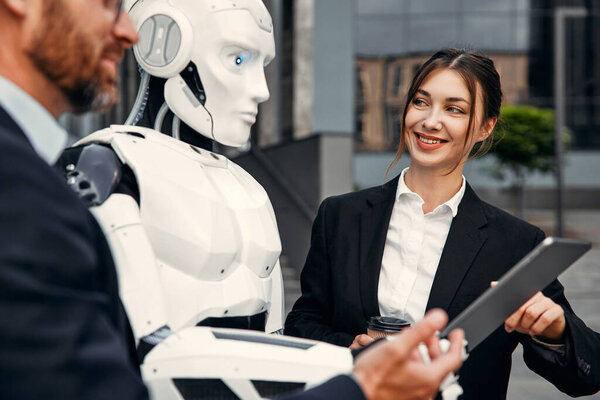 Young man and woman business partners in suits with a tablet standing together with a robot and discussing work against the background of a building. The future with artificial intelligence.