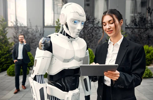 Takım elbiseli genç adam ve kadın iş ortakları ve tabletleri bir robotla yan yana duruyor ve bir binanın arka planında yapılan çalışmaları tartışıyorlar. Yapay zekayla gelecek.