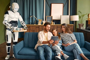 Oturma odasındaki kanepede kızıyla oturan bir aile tablet ve tepside çay ve kahve hazırlayan bir robot. İnsanlar ve yapay zeka arasında birlikte yaşamak.