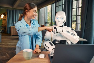 Elinde bir fincan kahve ve modern bir ofiste dizüstü bilgisayarın başında oturan insansı bir robot olan genç bir kadın yumruk yumruğa hareket ediyor. İnsanlar ve yapay zeka arasındaki işbirliği.