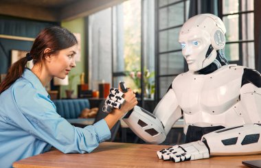 Genç bir kadın ve insansı robot modern bir ofiste masada otururken bilek güreşi yapıyorlar. İnsanlar ve yapay zeka arasındaki işbirliği.
